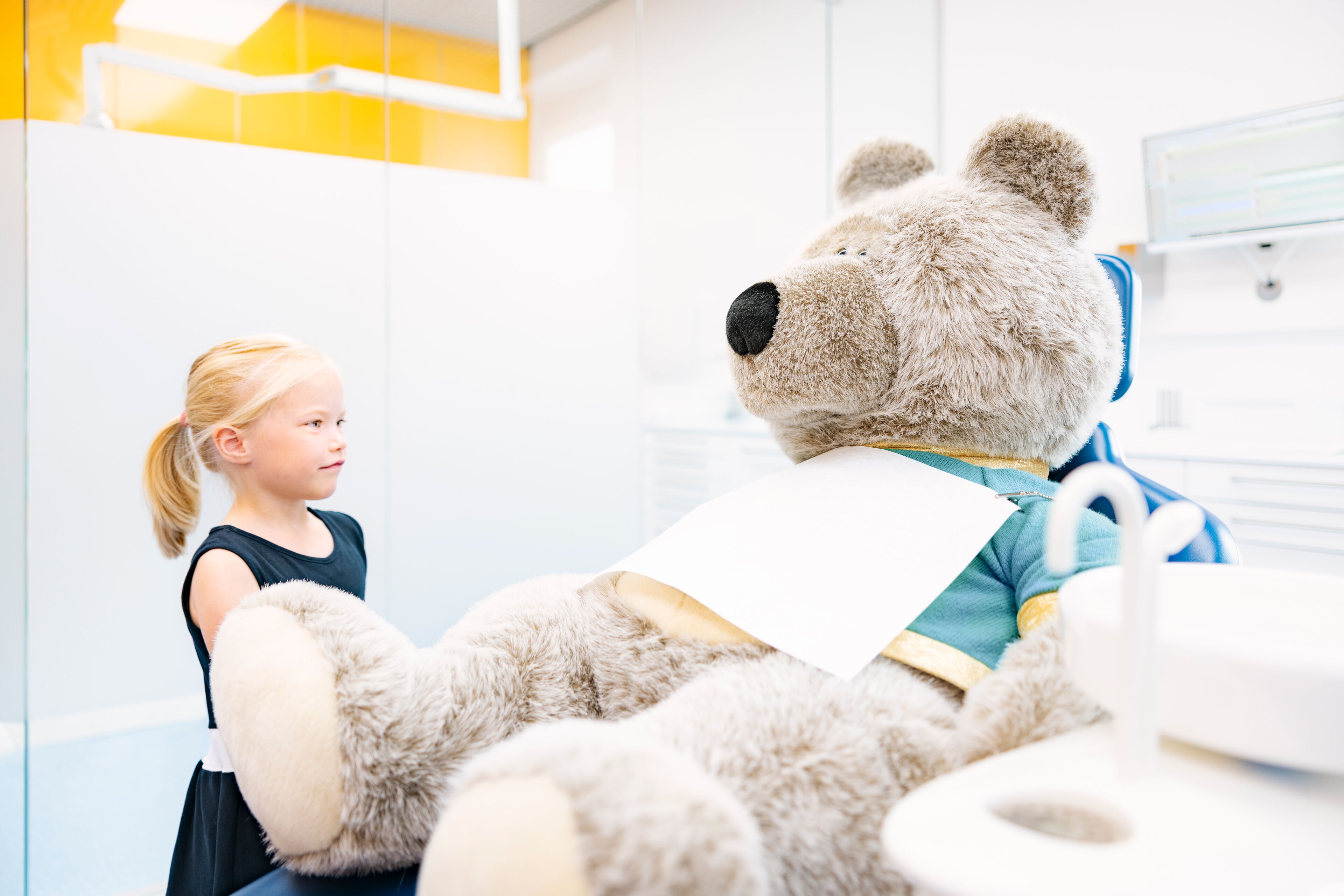 Kind in kieferorthopädischer Praxis schaut auf großen Plüschbären im Behandlungsstuhl